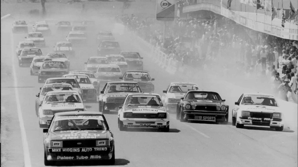 Carrera automovilística en blanco y negro con varios vehículos compitiendo en una pista llena de polvo y una multitud de espectadores en las gradas.