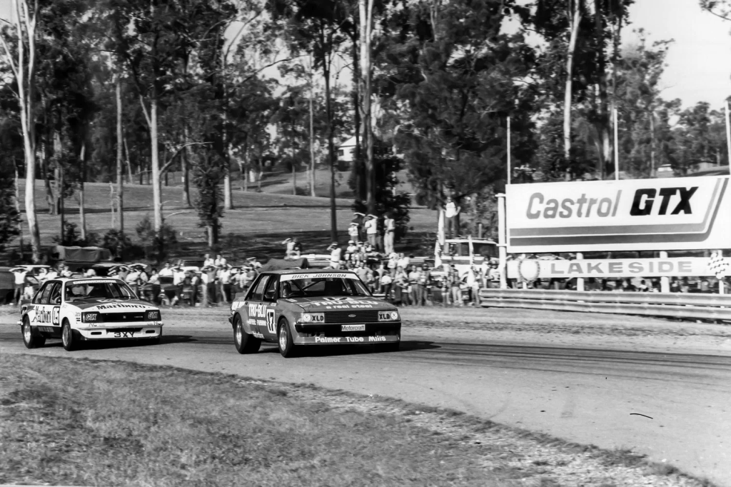 Dos coches de carreras compiten en una pista, rodeados de espectadores y árboles, con un cartel de Castrol GTX en el fondo.