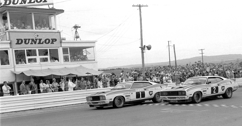 Dos autos de carrera cruzando la meta en una competición, con una multitud de espectadores y un edificio de control visible en el fondo.