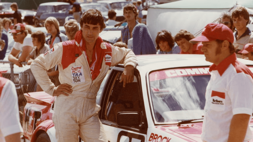 Piloto de carreras posando junto a un coche deportivo, con equipo de soporte y espectadores en el fondo.