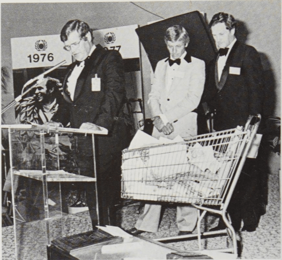 Un grupo de hombres de pie junto a un atril, uno de ellos hablando mientras los otros dos observan, con un carrito de compras lleno de documentos delante de ellos y un fondo que muestra años 1976 y 1977.