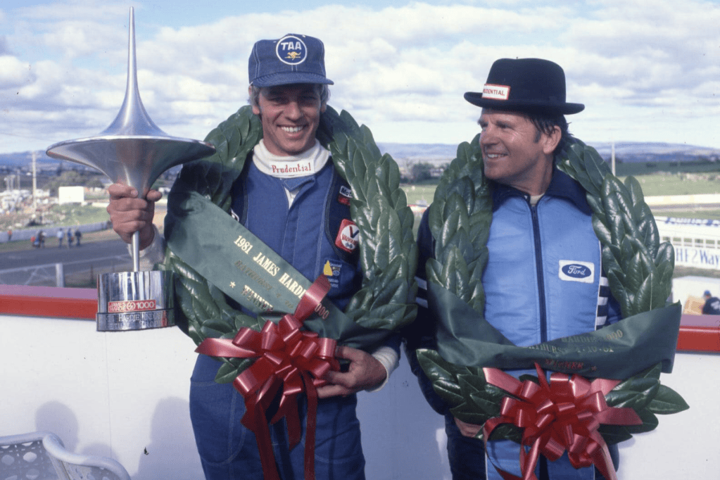 Dos pilotos de carreras sonrientes, uno sosteniendo un trofeo y ambos con coronas de laurel, en una ceremonia de premiación.